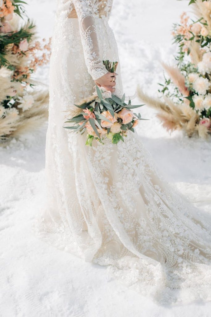 A bride in a lace gown holding a pastel bouquet in a snowy outdoor wedding.