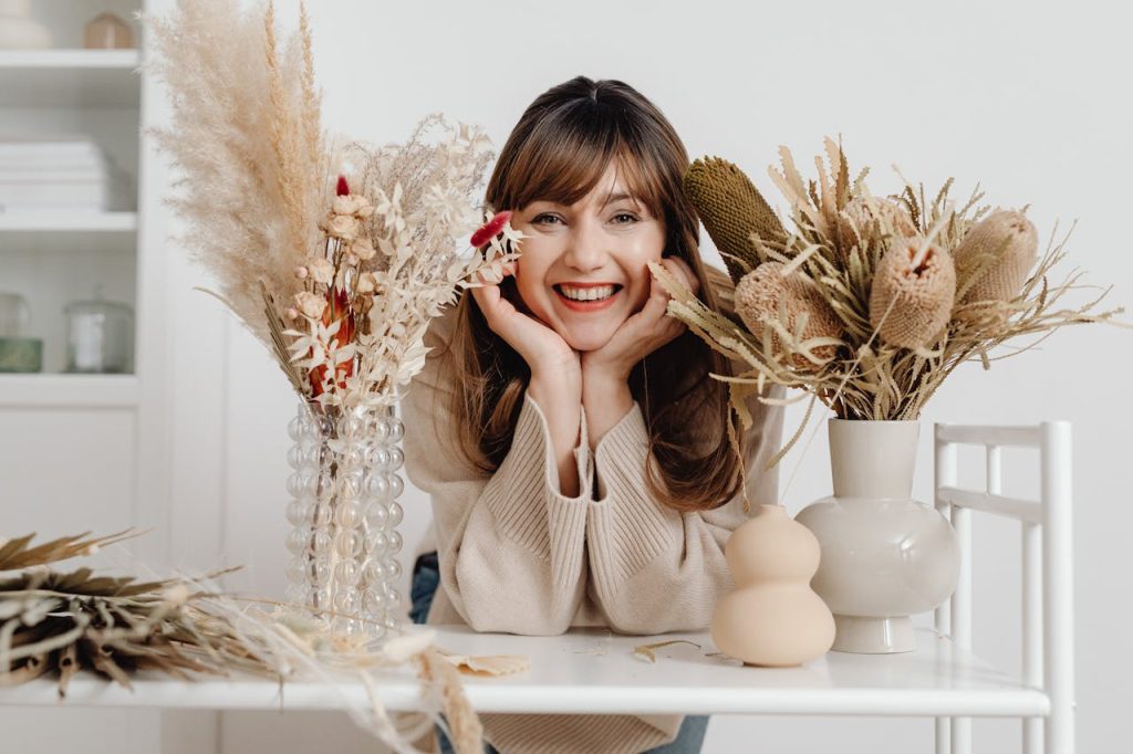 A cheerful woman seated indoors with beautiful dried flower arrangements on a table.